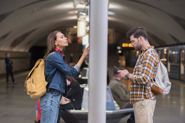 side view of female traveler with backpack looking at information board and boyfriend using smartphone at subway station