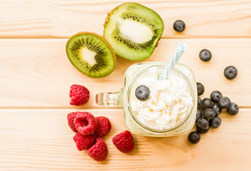 banana and kiwi milkshake in mason jar with creme on top and drinking straw decorated with fresh raspberries, blueberries and kiwi. flat lay top view smoothie on wooden background