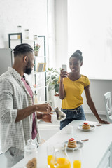 african american girlfriend taking photo of boyfriend with coffee with smartphone at kitchen