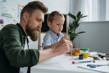 Naklejka premium side view of father and cute little son with paints and brushes drawing pictures together at home
