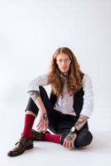 Fototapeta premium stylish tattooed businessman with curly hair sitting on floor and looking at camera on white