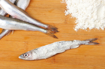 Raw smelt on a wooden board. There is flour beside it. One fish is covered in flour. Close-up. View from above. Light background.