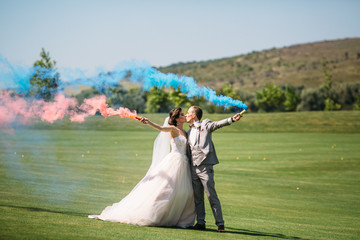 The bride and groom with smoke bombs on a field with green grass. Newlyweds walking outdoors at wedding day. Girl in luxury long white dress and man in business grey suit.
