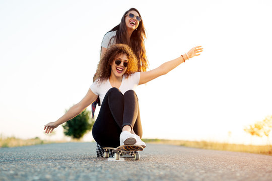 Happy young girl sitting on longboard being pushed by her friend
