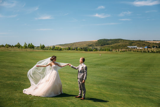 The newlyweds are walking along the golf course with green grass. The groom holds the bride's hand. Beautiful couple on wedding day for a walk. Girl in luxury long white dress with veil, man in