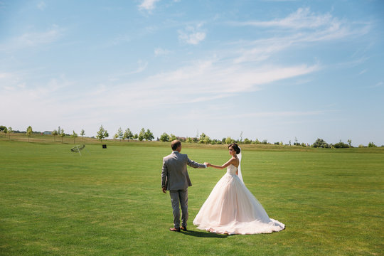 The newlyweds are walking along the golf course with green grass. The groom holds the bride's hand. Beautiful couple on wedding day for a walk. Girl in luxury long white dress with veil, man in