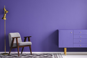 Golden lamp near a gray armchair and ultra violet cupboard in simple living room interior with copy...