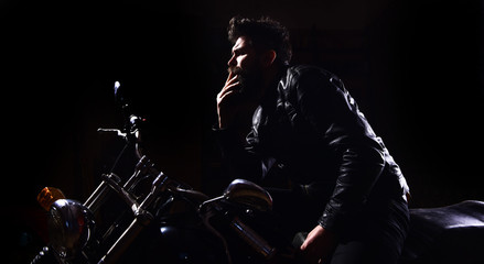 Fototapeta na wymiar Night racer concept. Man with beard, biker in leather jacket sitting on motor bike in darkness, black background. Macho, brutal biker in leather jacket riding motorcycle at night time, copy space.