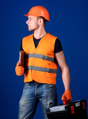 Man in helmet, hard hat holds toolbox and folder with documents, blue background. Repairman ready to work. Repair service concept. Worker, repairer, builder on dreamy face carries case with tools.