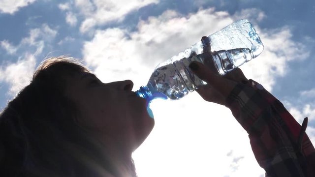 Young woman drinks a bottle of clean pure water on hot sunny day against sky background