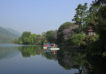 Green shore of lake Fewa, Pokhara. Small hindu temple and flowering trees. Boat.
