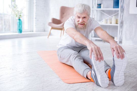 Supple Body. Upbeat Senior Man Bending Forward And Touching His Toes While Sitting On The Yoga Mat And Smiling At The Camera