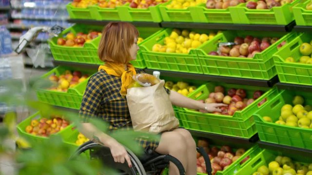 Cheerful woman with paralyzed legs riding wheelchair along produce aisle in supermarket and choosing apples