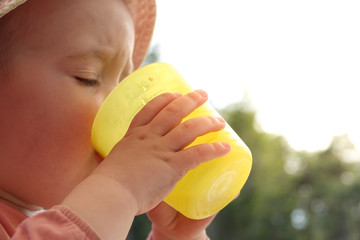 A little baby girl drinks water from a plastic glass. Thirsty child with sun hat under the sun a...
