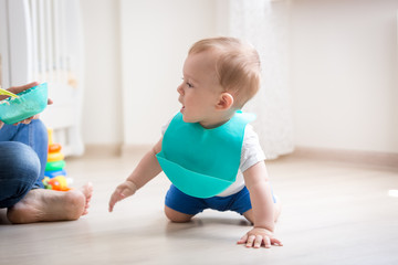 Cute toddler boy in apron crawling on floor while mother trying to feed him