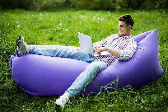 Handsome young man lying on inflatable sofa lamzac sufring in internet on laptop while resting on grass in park