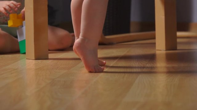 barefoot toddler on tiptoe near an armchair with kid playing on the background