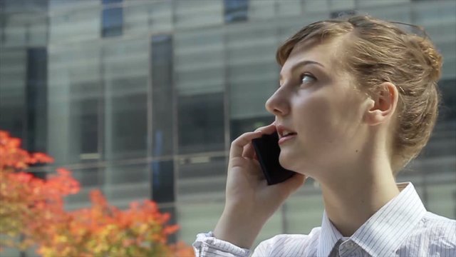 Business woman talking by phone in front of modern office buildings.