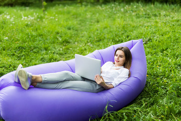 Pretty young woman lying on inflatable sofa lamzac use laptop while resting on grass in park