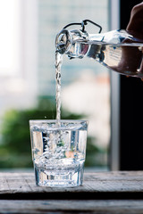 Clean drinking water is poured from a jug into a round glass cup on a wooden table and a light green napkin close-up macro on a green nature outdoors background .