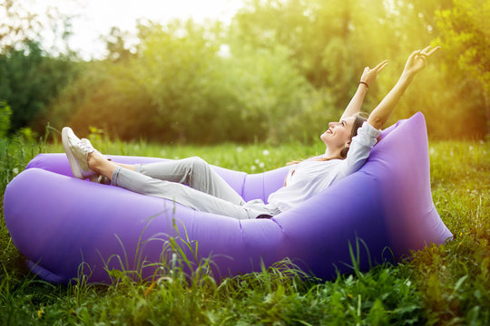 I am free. Pretty young woman lying on inflatable sofa lamzac with reised hands in the air while resting on grass in park