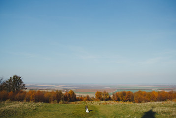 Fototapeta premium Wedding couple is walking in sunny day. Groom and bride in white satin lace dress are kissing with view on green and yellow field. Autumn nature panoramic landscape.