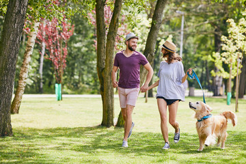 Lovely young couple running in the park with their dog during summer - obrazy, fototapety, plakaty