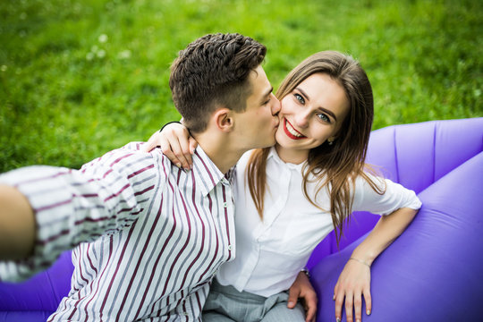 Young in love couple kissing while sitting on inflatable sofa lamzac and take selfie on phone resting on grass in park