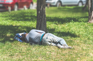 Old barefoot homeless or refugee man sleeping on the grass in the city park using his travel bag as pillow, social documentary street concept