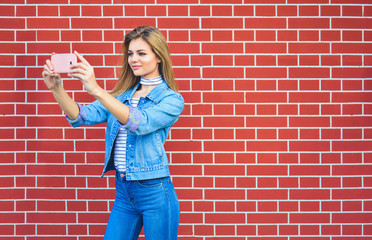 Young pretty woman taking selfie outdoors - Female summer fashion portrait - Teenager student holding mobile phone for selfi photo next to brick wall background
