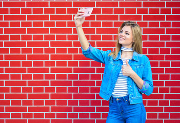 Young pretty woman taking selfie outdoors - Female summer fashion portrait - Teenager student holding mobile phone for selfi photo next to brick wall background