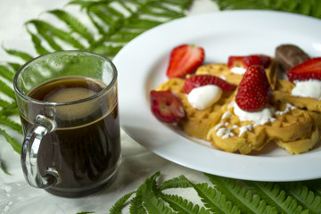 Waffles with strawberry, a cup of coffee and green leaves of fern on a table. Beautiful and tasty breakfast.