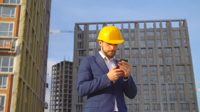 young attractive man in a yellow helmet makes photos of buildings under construction on a mobile phone. the person sends messages to the customer via a smartphone. 4k
