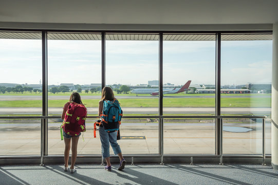 Two young women backpacking, waiting for plane at the airport to travel on holiday. Back side of traveler girl looking at the flying plane. travel and active lifestyle concept