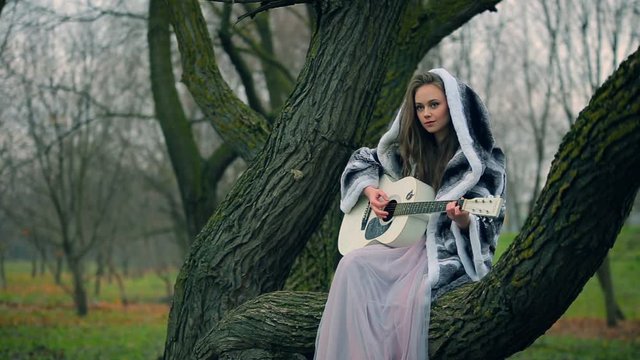 Caucasian teen girl pays guitar sitting on a tree in fairytale forest