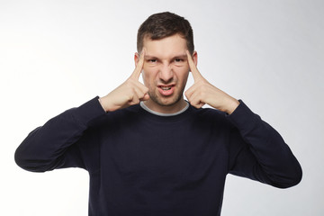 Headshot of young man holds fingers on temples, has bad memory, tries to concentrate,remember details of accident, presses teeth with irritation. Overworked man has headache, looks tired, aggressive.