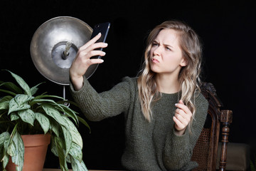 Unconfident student lady with blonde wavy hair taking pictures with smart phone, making faces at the camera. Young office female manager in bad mood making selfie using electronic device indoor.