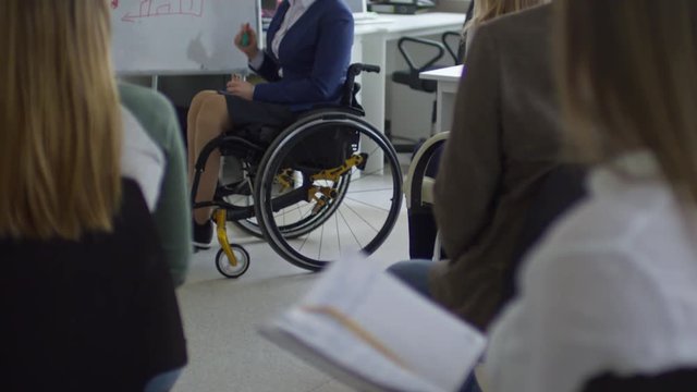 Tilt up of female business coach sitting in wheelchair and explaining bar graph on flipchart to group of office workers