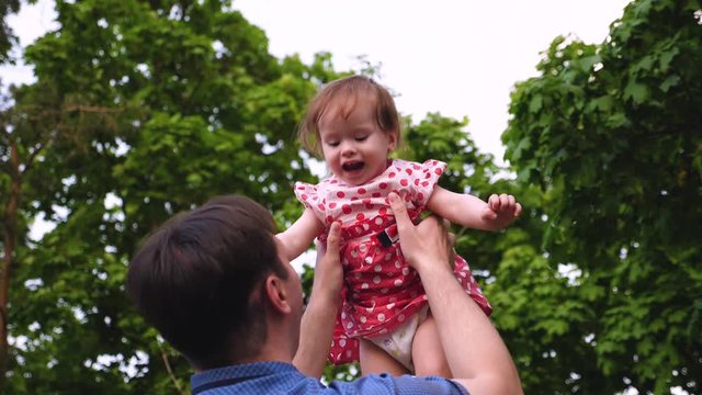 Baby laughs when his beautiful young dad pumps it through air. Dad is playing with baby in park for walk and smiling. Slow motion.