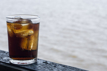 Glass of cola with ice on wooden table., with blur river outdoor background., hot concept. thirsty concept.