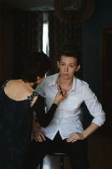 Portrait of beautiful sexy edgy young Caucasian woman sitting on chair indoors looking in camera. Makeup artist applying powder on model before shoot. Backstage candid shot of working people