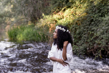portrait outdoors of a beautiful young afro american woman at sunset. Green background. Lifestyle at nature. Wearing white casual summer dress. She is standing in a river.