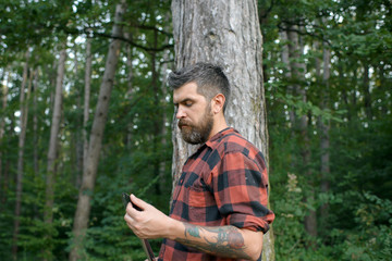 Tourist in plaid shirt travel with shovel. Bearded man in forest. Hipster with long beard on natural green landscape. Traveler hiking on sunny day. Summer vacation concept