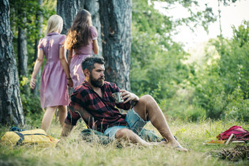 Bearded man drinking coffee or tea by campfire. Two girls in retro dresses walking on path in woods, turn back. Relaxation and tranquility concepts