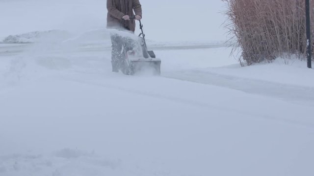 Wide shot of man pushing snow blower from the front, shooting snow to the side. Slow motion (120fps at 24)