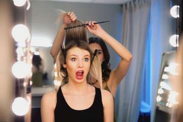 Hairstylist combs client's light-brown with blonde hair. Creating a luxurious evening hairstyle with pain. Dissatisfied customer unhaooy face.