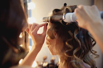 Gordijnen Hairstylist make beautiful curls with curler on light-brown with blonde client's hair. Creating a luxurious evening hairstyle with ringlets of medium length. © yolya_ilyasova