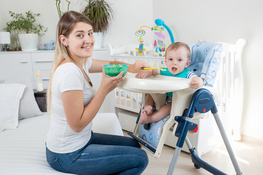 Portrait of smiling young mother feeding her toddler sin in highchair and looking in camera