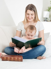Happy young mother reading big old book to her baby boy ion bed