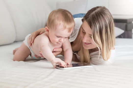 Cute baby boy in diapers lying on bed with mother and using digital tablet
